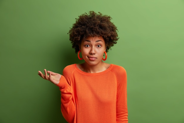 Doubtful indecisive woman raises palm with hesitation, faces difficult question or two choices, wears orange sweater and earrings, isolated on green wall. People, perception and attitude