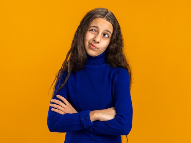 Doubtful teenage girl standing with closed posture looking up isolated on orange wall with copy space