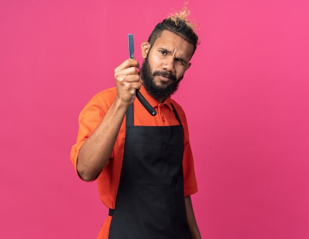 Doubtful young afro-american male barber wearing uniform showing straight razor 