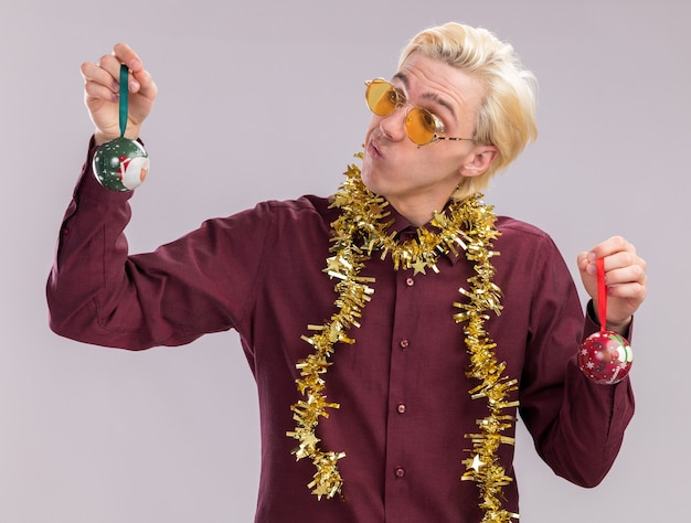 Doubtful young blonde man wearing glasses with tinsel garland around neck holding christmas baubles looking at one isolated on white wall