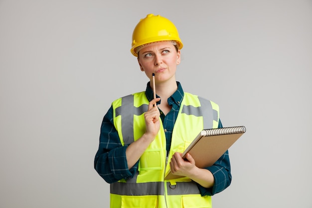 Doubtful young female construction worker wearing safety helmet and safety vest holding notepad touching chin with pencil looking up 