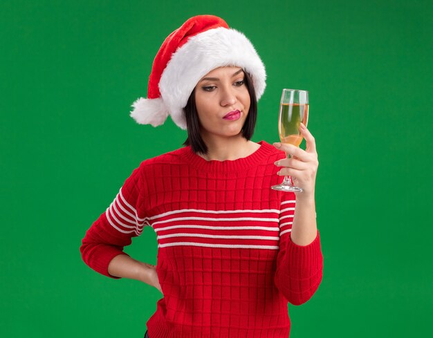 Doubtful young girl wearing santa hat holding and looking at glass of champagne keeping hand on waist isolated on green wall