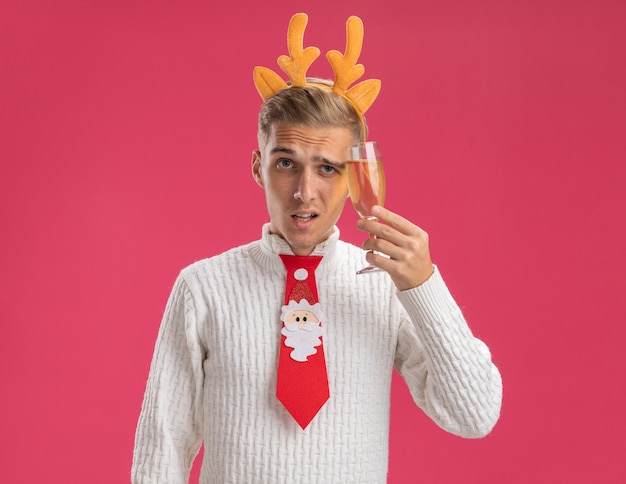 Free photo doubtful young handsome guy wearing reindeer antlers headband and santa claus tie holding glass of champagne touching head with it looking at camera isolated on pink background