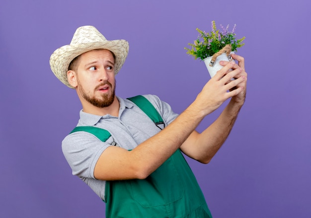 Doubtful young handsome slavic gardener in uniform and hat holding and looking at flowerpot isolated on purple wall