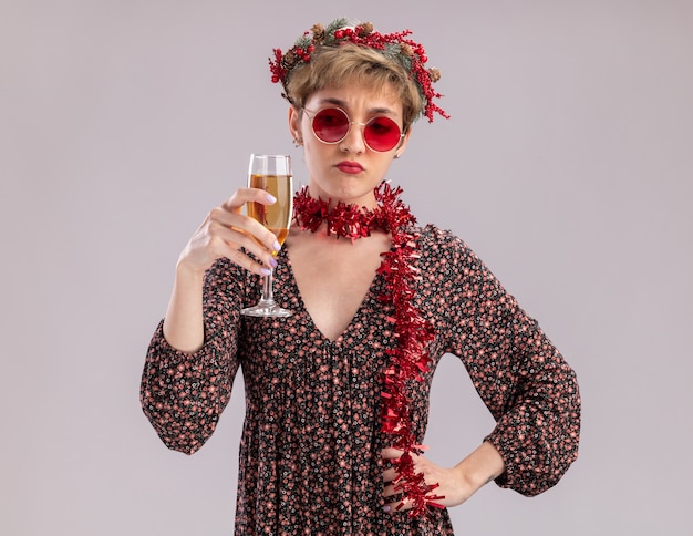 Doubtful young pretty girl wearing christmas head wreath and tinsel garland around neck with glasses holding and looking at glass of champagne keeping hand on waist isolated on white background