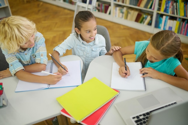 Draw attention. Top view of smiling girl touching hand of long-haired girlfriend writing and blonde boy sitting at table in library doing homework