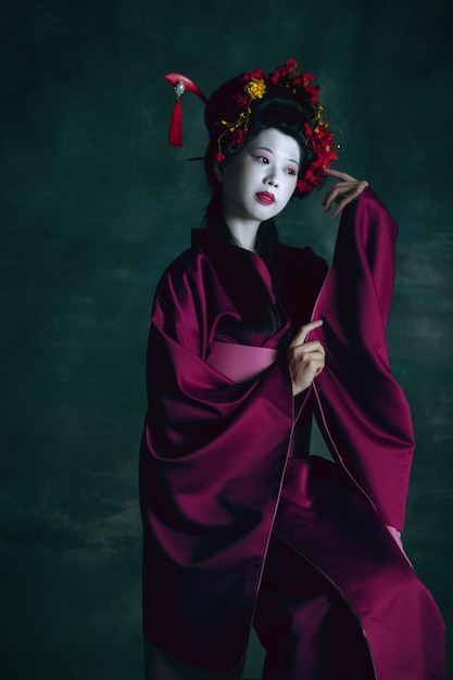 Free photo dreamful. young japanese woman as geisha isolated on dark green wall. retro style, comparison of eras concept. beautiful female model like bright historical character, old-fashioned.