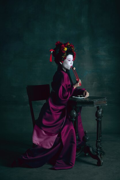 Dreamful. Young japanese woman as geisha isolated on dark green wall. Retro style, comparison of eras concept. Beautiful female model like bright historical character, old-fashioned.