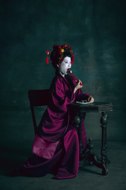 Dreamful. Young japanese woman as geisha isolated on dark green wall. Retro style, comparison of eras concept. Beautiful female model like bright historical character, old-fashioned.