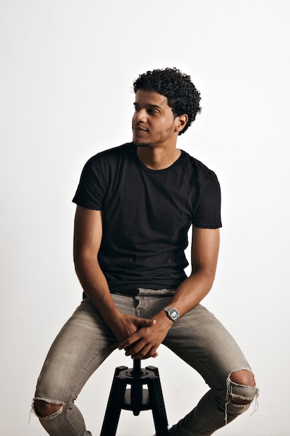 Dreamy athletic relaxed black young man wearing torn jeans and an unlabeled black t-shirt sitting on small stool on white wall