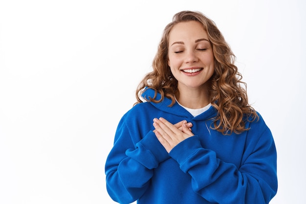 Free photo dreamy beautiful woman remember lovely heartfelt moment, holding hands on chest and smile with closed eyes, daydreaming, thinking about something, white wall