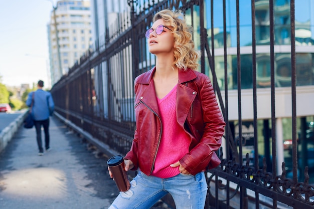 Dreamy blond girl walking the streets, drinking coffee or cappuccino. Stylish autumn outfit, leather jacket and knitted sweater. Pink sunglasses.