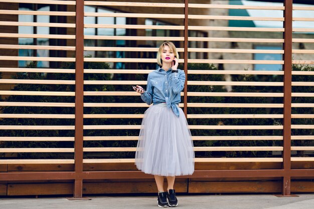 Dreamy blonde girl with bright pink lips standing and listening to music on a smartphone with striped wooden balks behind 