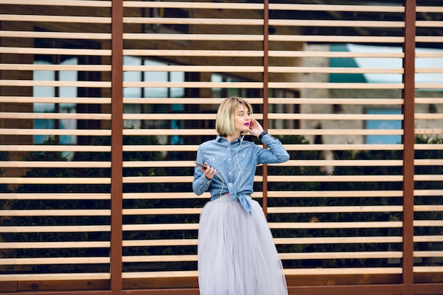 Dreamy blonde girl with bright pink lips standing and listening to music on a smartphone with striped wooden balks behind 