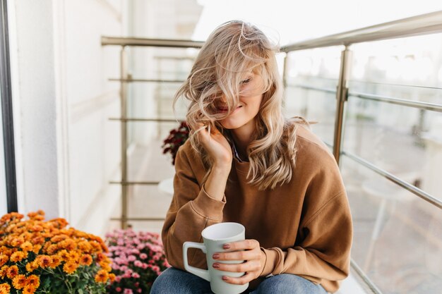 Dreamy blonde woman sitting at balcony with cup of tea. Stunning female model chilling at terrace.