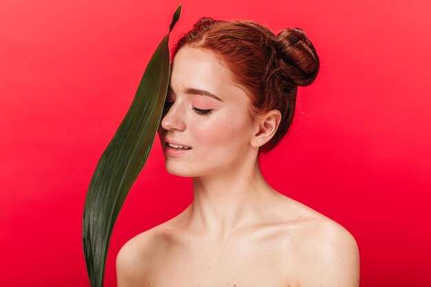 Dreamy caucasian girl with green leaf smiling on red background Studio shot of attractive nude ginger woman with plant