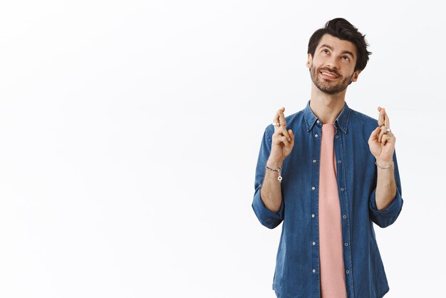 Dreamy charming bearded 25s guy in hipster shirt tshirt looking up daydreaming or fantasizing cross fingers for good luck making wish smiling thoughtful imaging dream come true white background