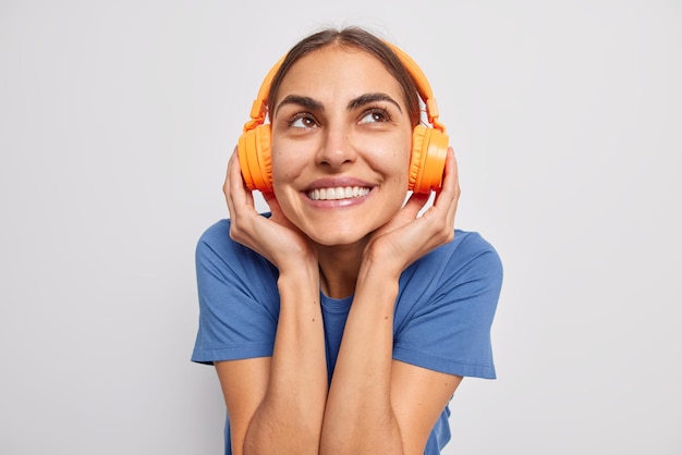 Free photo dreamy cheerful young woman keeps hands on wireless headphones smiles pleasantly enjoys favorite song dressed in casual blue t shirt isolated over white background. music beat for energetic mood