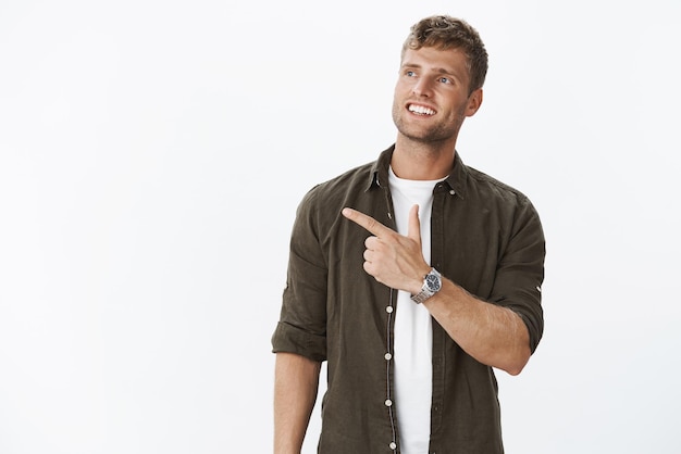 Free photo dreamy and delighted successful lucky guy with fair hair blue eyes and white teeth pointing at upper left corner and looking impressed and fascinated smiling from satisfaction against grey wall