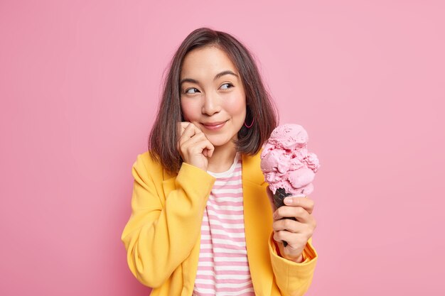 Dreamy eastern woman looks pleasantly away with thoughtful expression holds big tasty ice cream thinks about something keeps hand on face dressed in stylish clothes isolated on pink wall