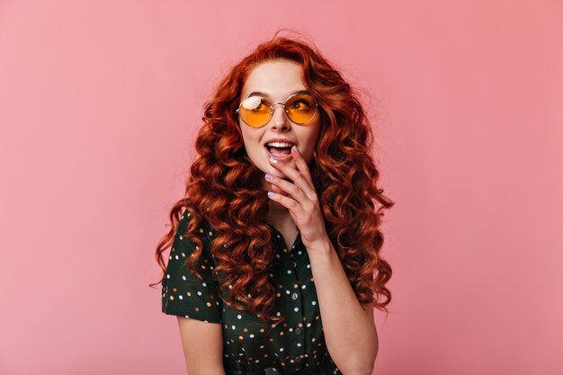 Dreamy ginger girl looking away with open mouth. Studio shot of emotional young woman in sunglasses posing on pink background.