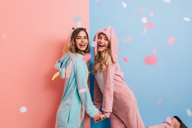 Dreamy girl in rabbit kigurumi holding hands with friend Ecstatic ladies in cute pajamas dancing and smiling