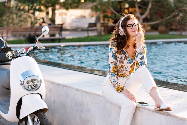 Free photo dreamy girl with elegant hairstyle listening music and looking away, spending time near fountain