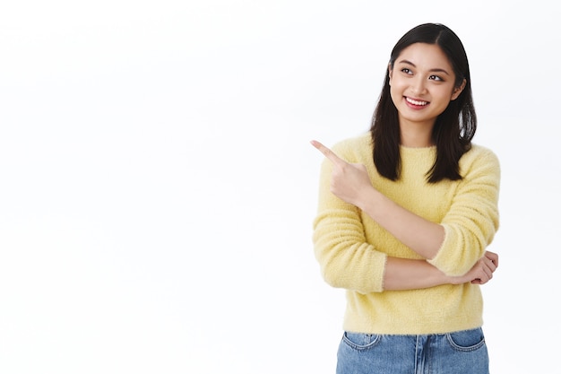 Free Photo dreamy happy asian girl with beaming white smile, pointing finger left and looking satisfied at advertisement, found excellent choice, picking product, making decision, ready order, white wall