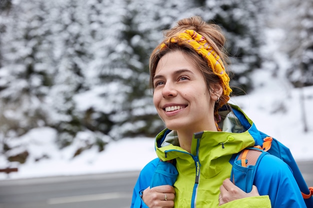 Dreamy happy woman with cheerful expression, wears yellow headscarf and anorak, carries rucksack