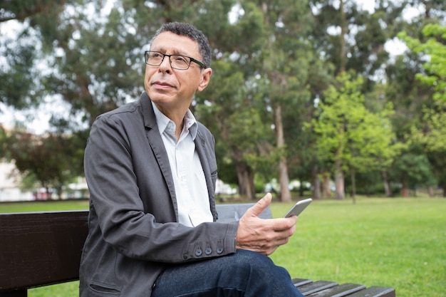 Dreamy man using smartphone and sitting on bench in park