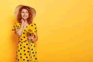 Free photo dreamy redhead woman posing in yellow polka dress and straw hat