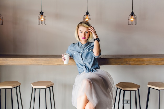 Dreamy stylish girl with blonde hair and pink lips sitting in a coffee shop with wooden chairs and table. She holds a cup of cofee and touches her hair