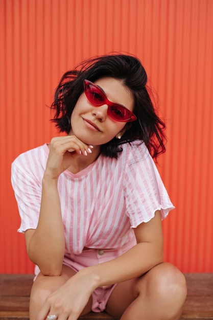 Free photo dreamy tanned woman propping face with hand and looking at camera. outdoor shot of smiling woman in pink sunglasses.