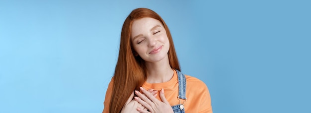 Dreamy touched romantic ginger girlfriend close eyes recalling heartwarming romance touch heart palms pressed chest smiling tenderly feel love care sympathy express affection blue background