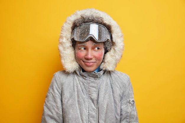 Free Photo dreamy winter girl with red frozen face enjoys holiday mountain resort during cold good snow day covered by snowflakes dressed in warm jacket with hood wears ski goggles likes extreme sport.