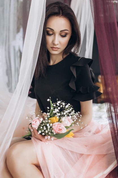 Free Photo dreamy woman in black dress sits on pink bed and holds bouquet on her knees