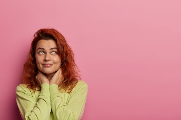 Free photo dreamy woman in love recalls perfect date with boyfriend, thinks about cute sweet things, keeps hands on neck, looks somewhere, wears green sweater