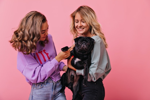 Free photo dreamy women playing with cute bulldog puppy on pastel. romantic girls enjoying good day and posing with pet.