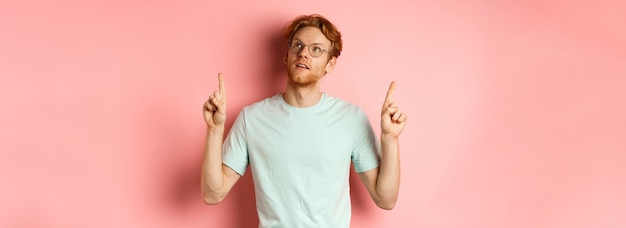 Free photo dreamy young man with red hair and beard pointing fingers up looking pensive at top standing against