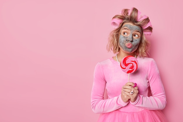 Free Photo dreamy young woman keeps lips folded applies nourishing clay mask wears hair rollers for making hairstyle wears festive dress holds lollipop on stick isolated over pink background blank space