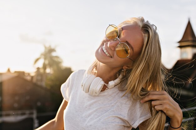 Dreamy young woman wears big white headphones posing on sky background with cute smile.