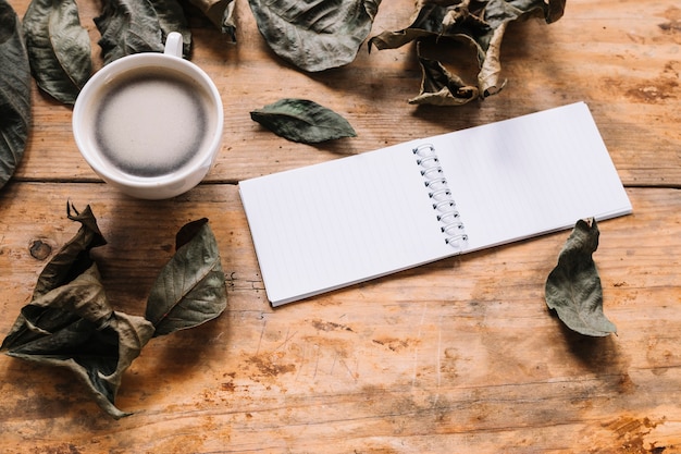 Dried leaves, cup of coffee and notebook on wooden background