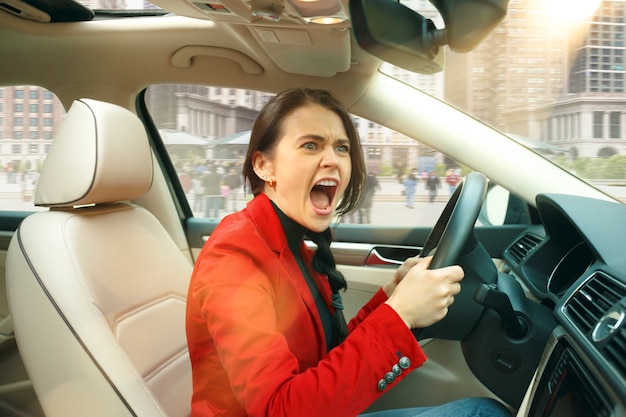 Free photo driving around city. young attractive woman driving a car. young pretty caucasian model in elegant stylish red jacket sitting at modern vehicle interior. businesswoman concept.