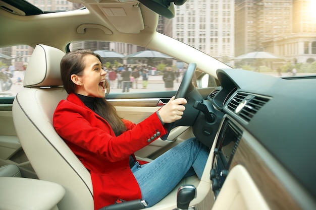 Driving around city. Young attractive woman driving a car. Young pretty caucasian model in elegant stylish red jacket sitting at modern vehicle interior. Businesswoman concept.