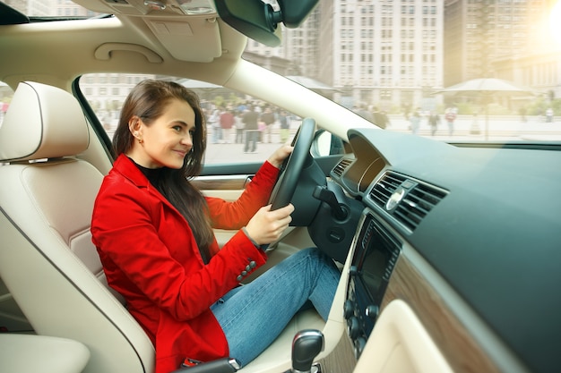 Driving around city. Young attractive woman driving a car. Young pretty caucasian model in elegant stylish red jacket sitting at modern vehicle interior.