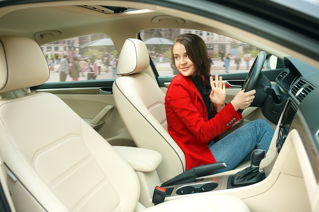 Free photo driving around city. young attractive woman driving a car. young pretty caucasian model in elegant stylish red jacket sitting at modern vehicle interior