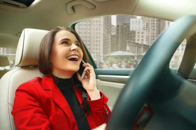 Driving around city. Young attractive woman driving a car. Young pretty caucasian model in elegant stylish red jacket sitting at modern vehicle interior