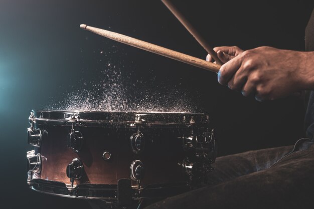 Drummer using drum sticks hitting snare drum with splashing water on black background under studio lighting close up.