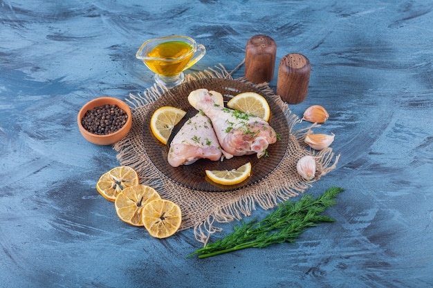 Free photo drumsticks and lemon on a glass plate next to dried lemon, spice bowl, dill, and oil on a burlap napkin on the blue surface