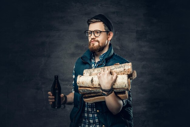 A drunk bearded male dressed in fleece shirt holds firewoods and craft bottled beer.